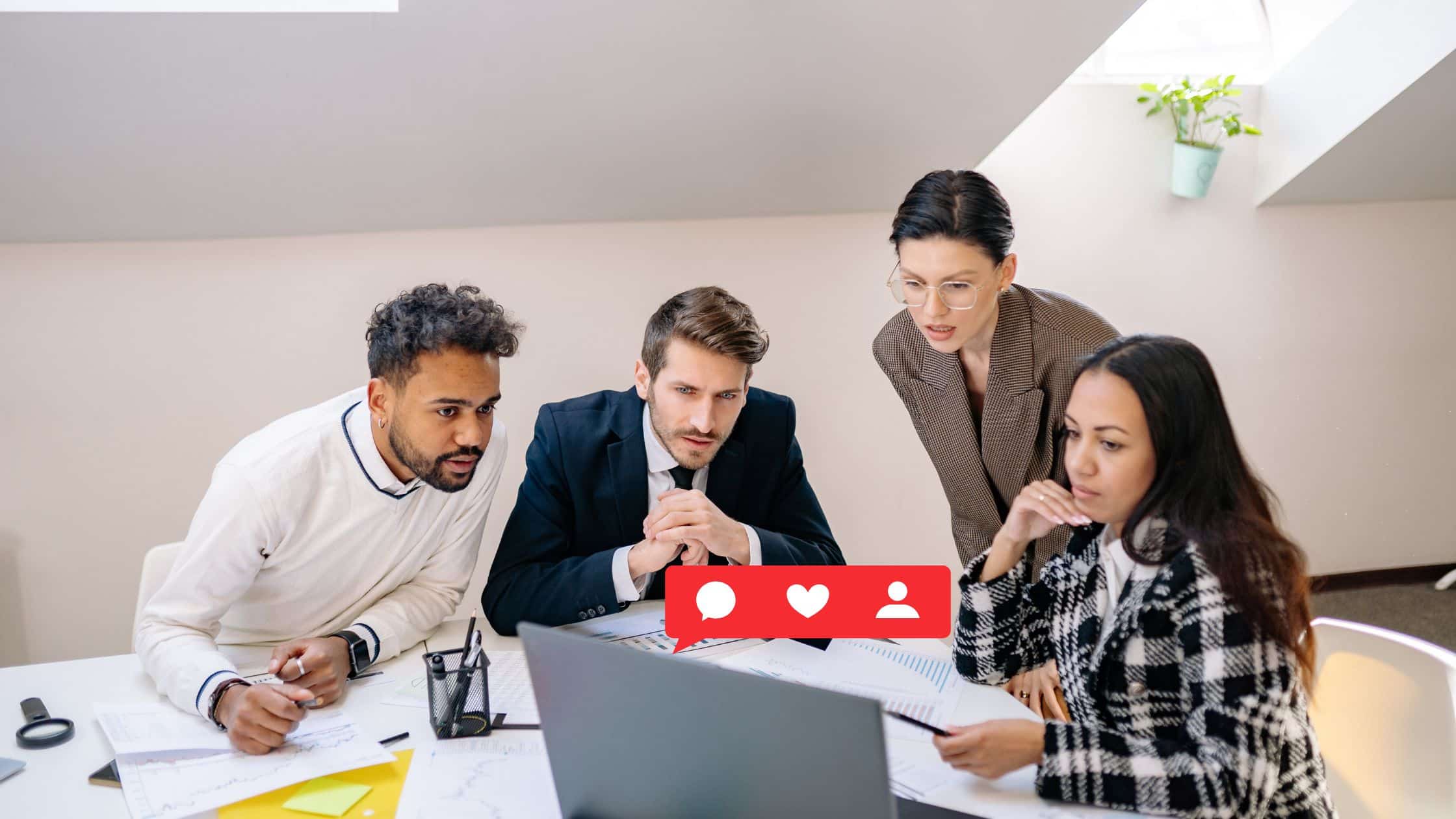 A group of four people in the top floor office looking at a laptop with a social media engagement icon popping up out of the laptop