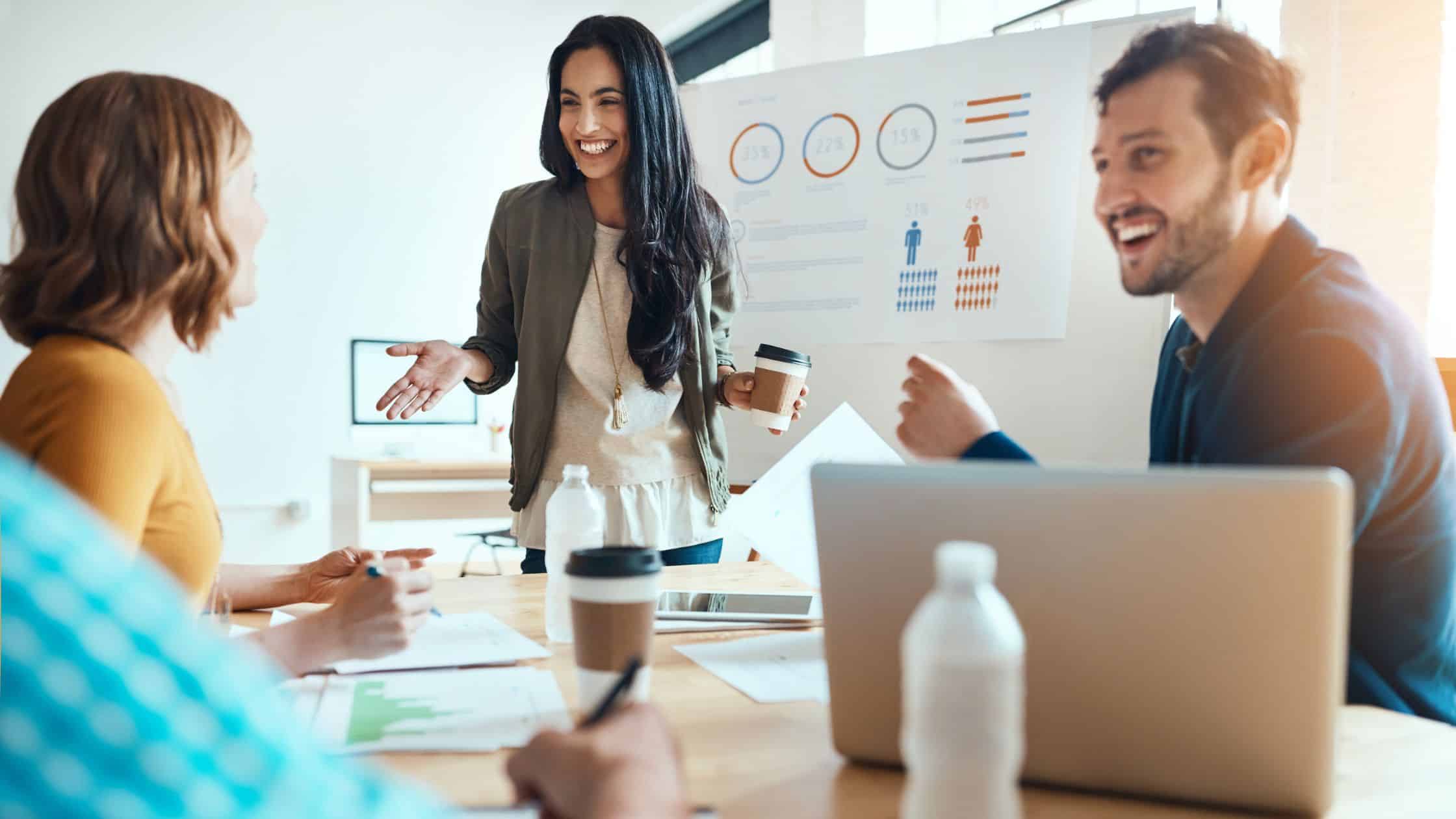 3 people sitting around a table in a office. One woman standing with a coffee in hand giving a presentation on the demographics of social media platforms on a large board behind her
